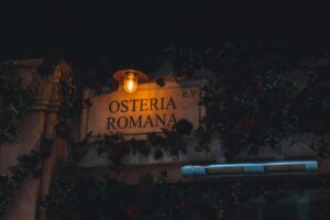 Warmly lit Osteria Romana sign surrounded by foliage, creating an inviting nighttime ambiance.